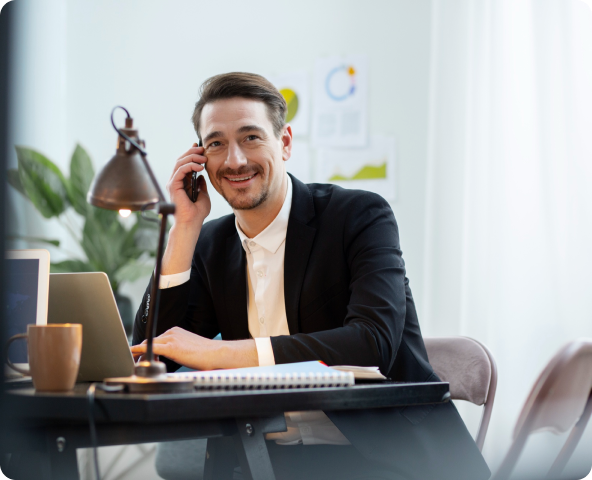 Homem de terno sentado em sua mesa e falando ao telefone com uma expressão de felicidade.